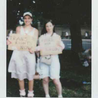 Two people holding protest signs stand together outdoors, one sign says "Trans Rights Now" and the other is too blurry to make out.