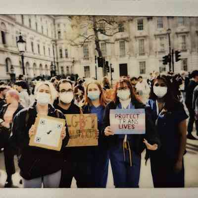 A group of people, wearing masks, hold signs saying phrases like "Protect Trans Lives".