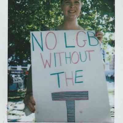 A person smiles while holding a sign reading, "No LGB Without the T," in a park
