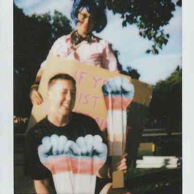 In the image, two people are outdoors holding signs with creative artwork, surrounded by trees.