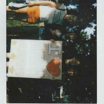 Three people are holding a large sign with love symbolism, standing outdoors on a sunny day.