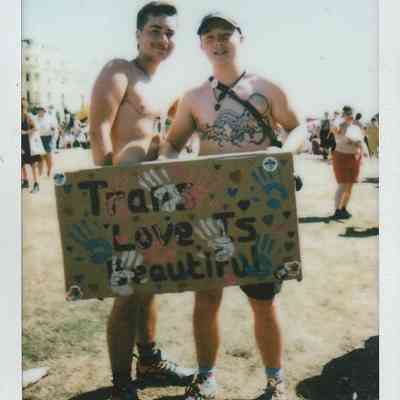 Two people smiling, holding a colorful sign that reads "Trans Love Is Beautiful".