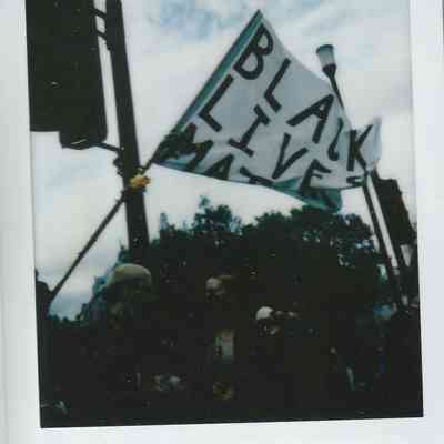 A group of people gathers under a "Black Lives Matter" flag, demonstrating in daylight.