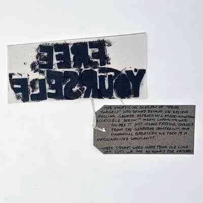 The image shows a reversed "FREE YOURSELF" lino cut stamp covered in black ink. With a brown, handwritten luggage tag.