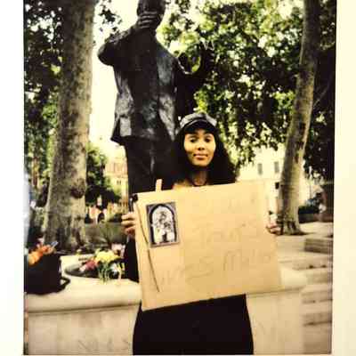 A person holds a sign reading "Black Trans Lives Matter" in front of a statue with trees behind.