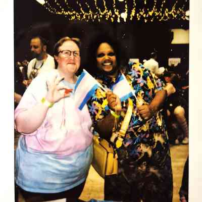 Two smiling individuals hold small transgender flags under decorative lights at a lively indoor event.
