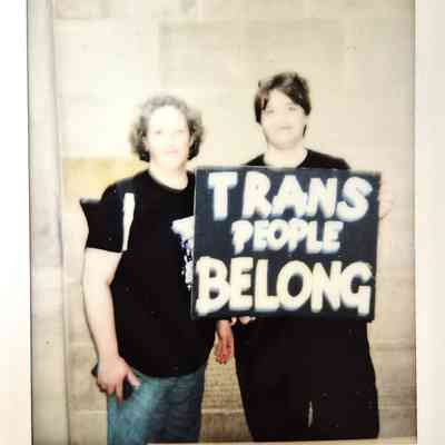 Two individuals, in black attire, hold a sign that says “Trans People Belong”.