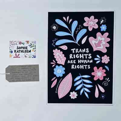 A print showing the text "TRANS RIGHTS ARE HUMAN RIGHTS" in white on a black background decorated with pink, white and blue leaves and flowers. With a name card, SOPHIE KATHLEEN, decorated with flowers, and a brown, handwritten luggage tag.