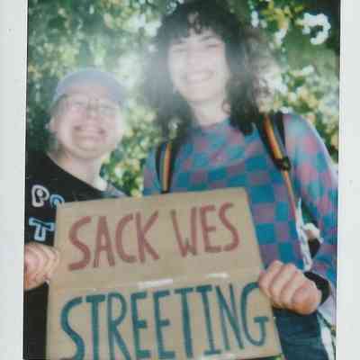 Two people are smiling and holding a "Sack Wes Streeting" sign, standing under leafy trees.