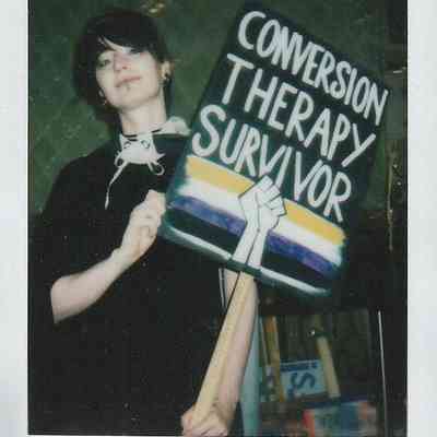 A person holds a sign reading "Conversion Therapy Survivor" with a rainbow flag design.