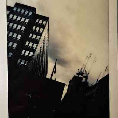 The image shows tall buildings and cranes silhouetted against an overcast sky, captured in a vintage photo