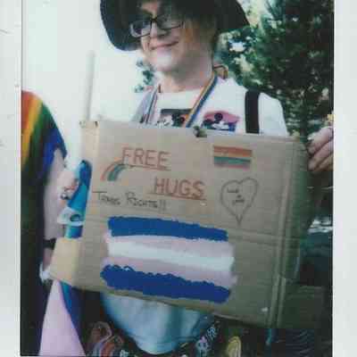 A person holds a "Free Hugs" wearing a hat and rainbow accessories.