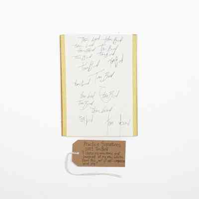 The image shows a sheet with handwritten practice signatures and a brown tag with writing on it sat underneath.