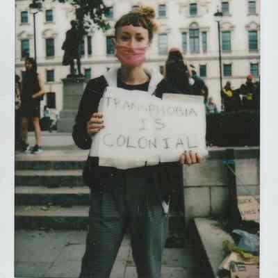A person in a pink mask holds a sign that says, "Transphobia is Colonial".