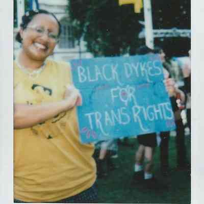 A person in a yellow shirt holds a sign which says "Black Dykes For Trans Rights".