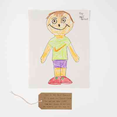 A child's crayon drawing of a smiling boy. He has brown hair and wears a green t-shirt with an orange tick, purple shorts and red shoes with a green spot. With a brown, handwritten luggage tag.