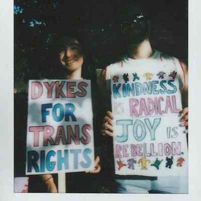 Two people hold signs saying "Dykes for Trans Rights" and "Kindness is Radical, Joy is Rebellion".
