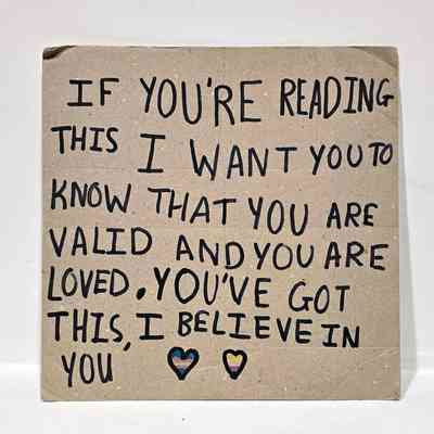Handwritten cardboard placard with text "IF YOU'RE READING THIS I WANT YOU TO KNOW THAT YOU ARE VALID AN YOU ARE LOVED. YOU'VE GOT THIS, I BELIEVE IN YOU" followed by two hearts coloured in the trans and non-binary colours.