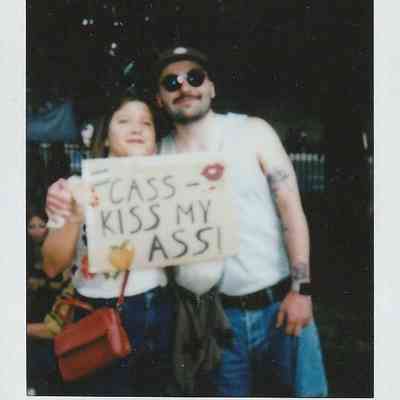 Two people stand together outdoors, holding a protest sign which says "CASS Can Kiss My Ass".
