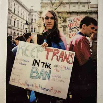 A person holds a sign which says "KEEP TRANS IN THE BAN" and various hashtags such as #TransRightsAreHumanRights.