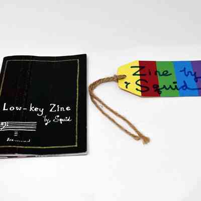 A black zine which says 'Low Key Zine by Squid' on the front. Next to the zine is a tag which is rainbow coloured and says in black marker 'Zine Squid'.