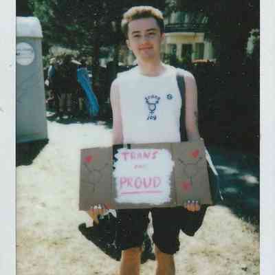 In a sunny outdoor setting, a person holds a "Trans and Proud" sign, smiling confidently.