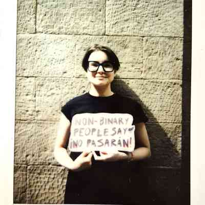 A person holds a sign reading, "Non-binary people say ¡No pasarán!" against a wall.