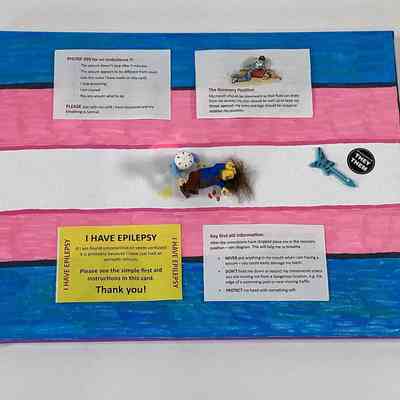 The image shows a canvas, colored in the the trans pride colours,  with epilepsy information, including emergency instructions. There are two Lego figures demonstrating the recovery position.