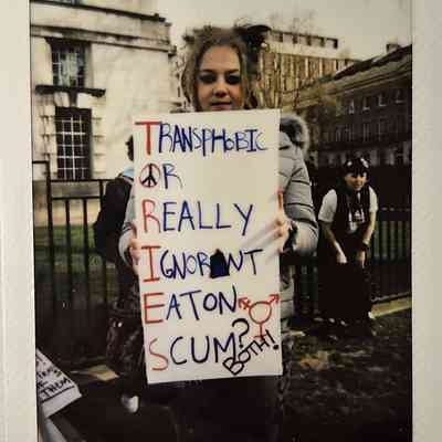 A person holds a protest sign which says "TRANSPHOBIC OR REALLY IGNORANT EATON SCUM? BOTH!"