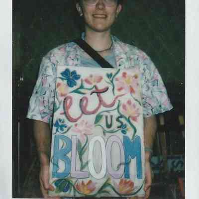 A person holding a colorful floral-themed sign reading "Let Us Bloom," smiling in a casual setting.