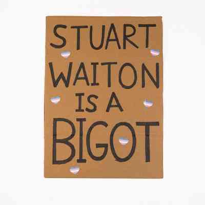 A placard whicch says 'Stuart Waiton is a Bigot' with small trans coloured heart stickers stuck on it.