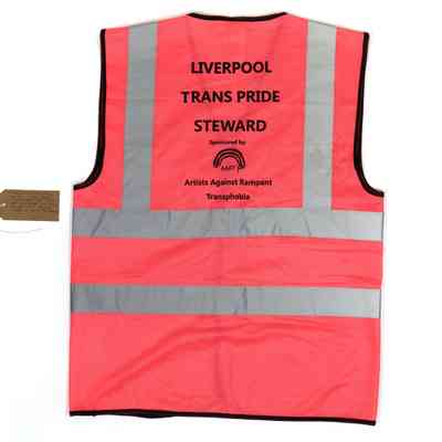 A pink Hi-Vis vest with the text "LIVERPOOL TRANS PRIDE STEWARD Sponsored by Artists Against Rampart Transphobia" printed in black on the back. With a brown, handwritten luggage tag.