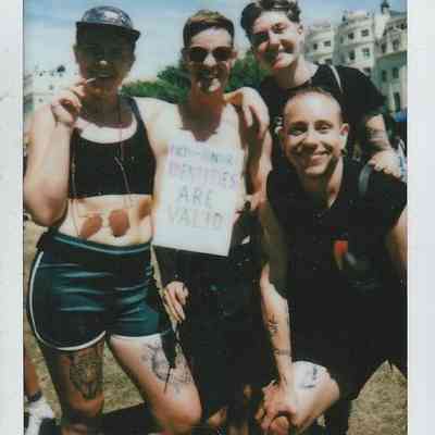 Four people are smiling outdoors, with one holding a sign saying "Non-binary identities are valid."