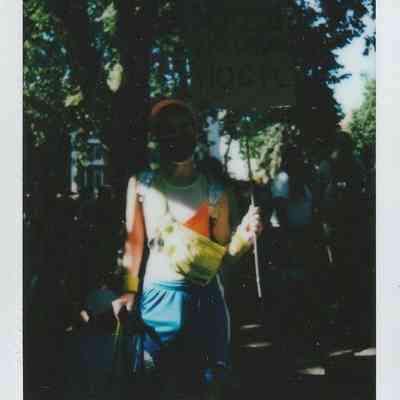 A person in bright clothing holds a sign amid a sunny outdoor setting with trees.