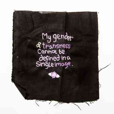 Piece of black cotton fabric with embroidered text in centre, square, frayed on all edges & unhemmed. Embroidered with mauve, purple & light green thread, reads; 'My gender & transness cannot be defined in a single image' with flower & leaves motif beneath