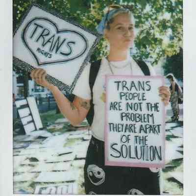A person holds two signs advocating trans rights, standing outside under trees with sunlight filtering through leaves.
