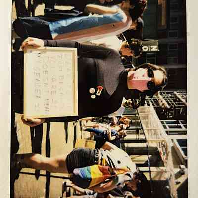 A person wearing a mask holds a sign at a busy outdoor protest or gathering event.