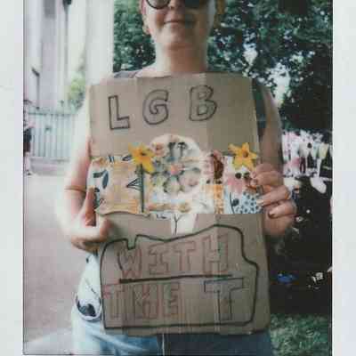 A person holds a cardboard sign reading "LGB with the T," decorated with flowers and stars.