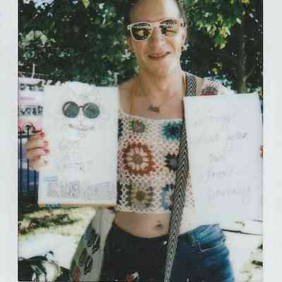 A person wearing sunglasses holds two signs advocating for trans rights while standing outdoors by trees.