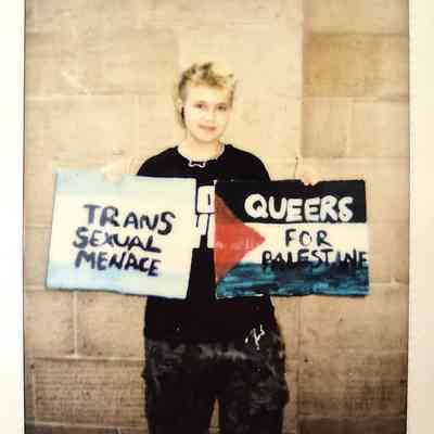 A person holds signs reading "Trans Sexual Menace" and "Queers for Palestine".