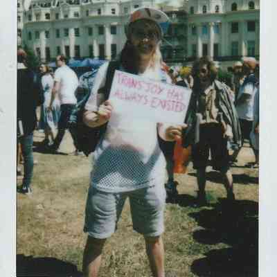A person stands outside holding a sign that says "Trans Joy Has Always Existed" in bright sunlight.
