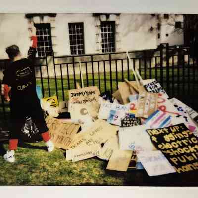 A person stands raising a fist beside a pile of protest signs on the grass, supporting trans rights.