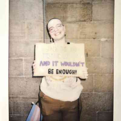 A person stands smiling against a stone wall, holding a placard.