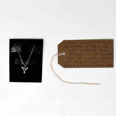 A necklace with the trans symbol displayed in a box with a brown luggage tag next to it.