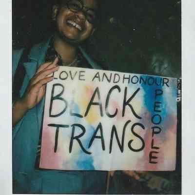 A person smiles while holding a colorful sign that says, "Love and Honour Black Trans People."