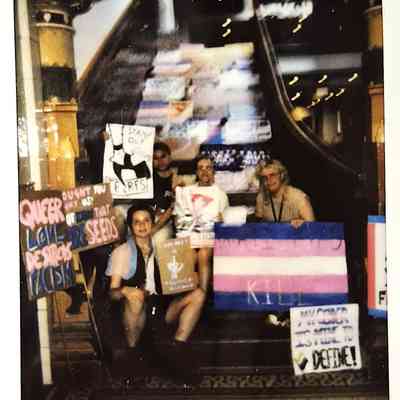 A group of people sits on stairs holding signs with messages supporting LGBTQ+ and social causes.
