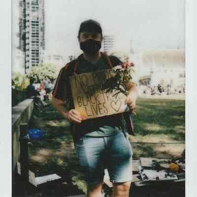 A person, wearing a mask, holds a sign which says "No Pride Without Black Trans Lives" standing in a park.