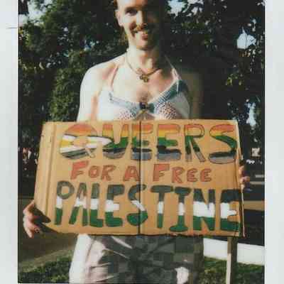 A person stands outdoors holding a sign reading "Queers for a Free Palestine" in colorful letters.