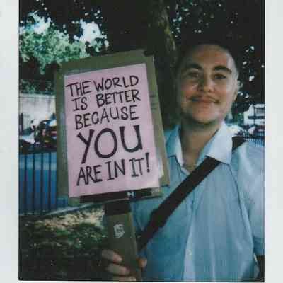 A person stands outside, smiling and holding a sign that reads, "The world is better because you are in it!"