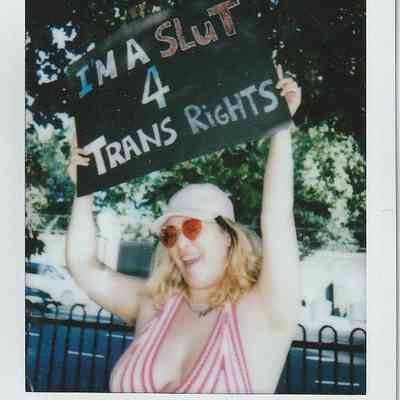 A person holds a sign supporting trans rights while smiling outdoors, wearing sunglasses and a striped top.